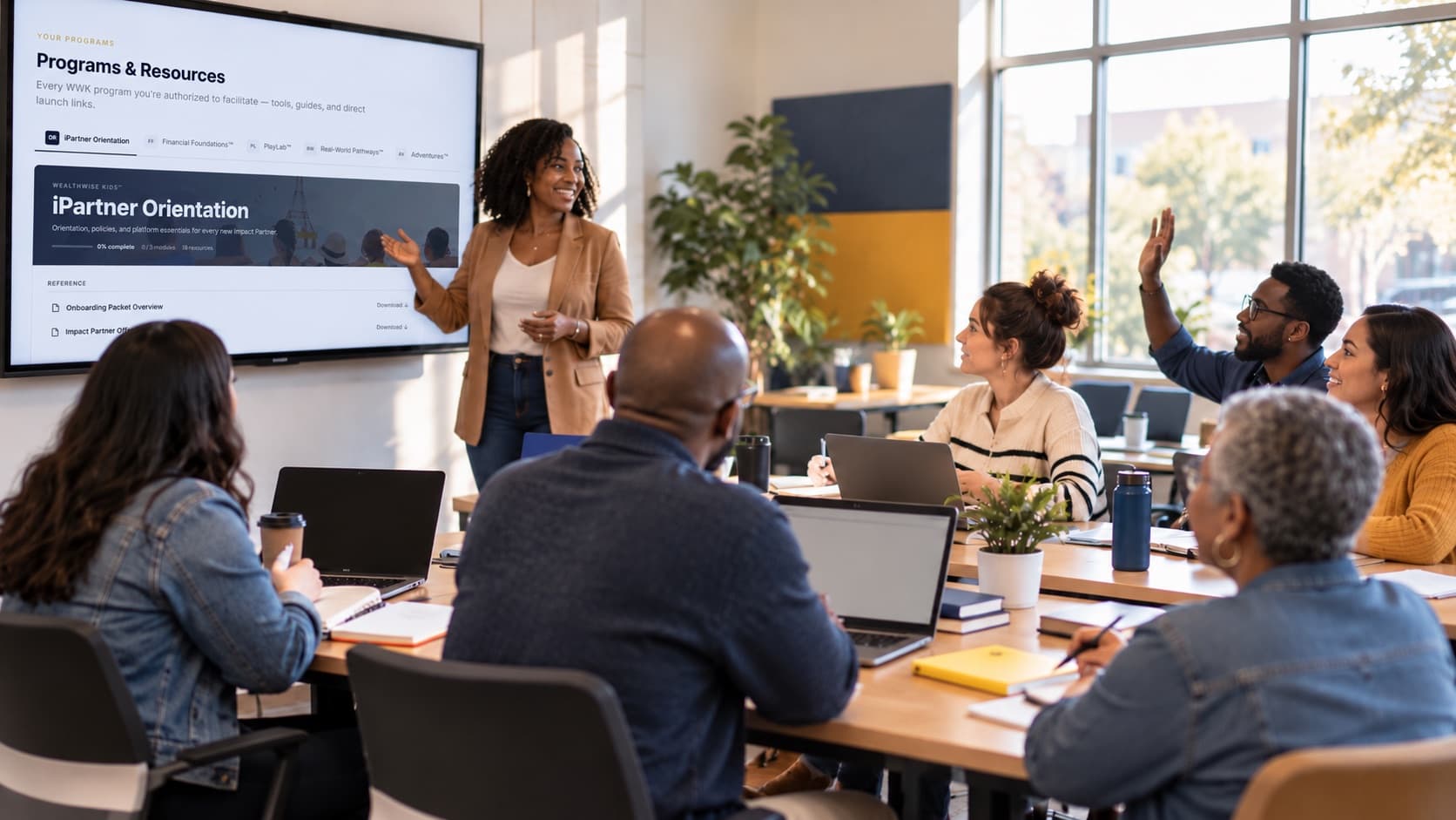 A trainer presents the iMPACT Partners portal's iPartner Orientation on a large wall screen to a cohort of adult educators at laptops; one raises a hand