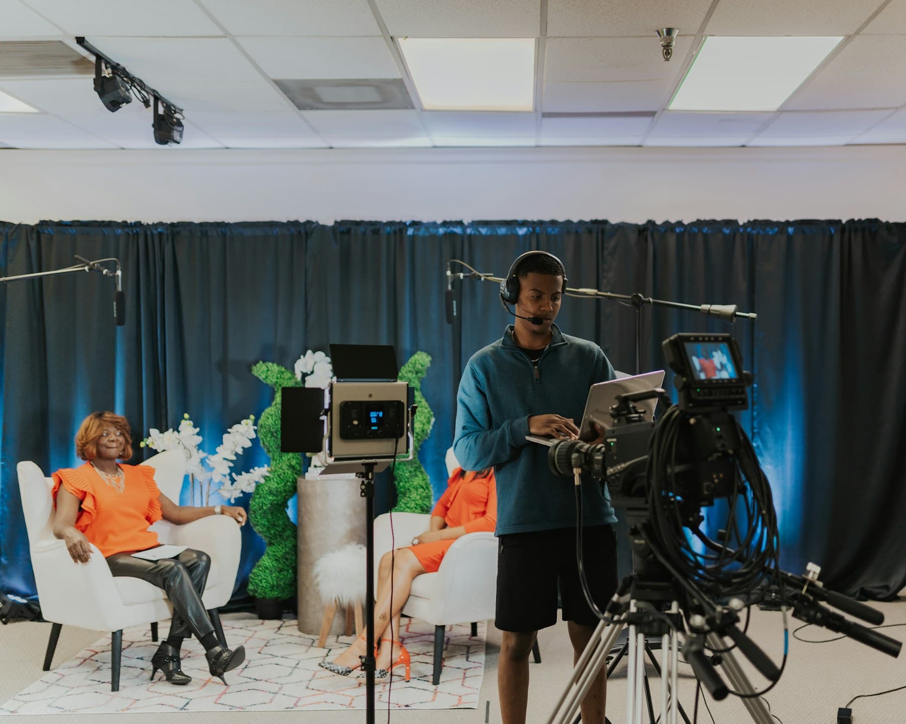 A bright interview set: two hosts seated in armchairs before a navy curtain, a camera operator and boom microphone in the foreground