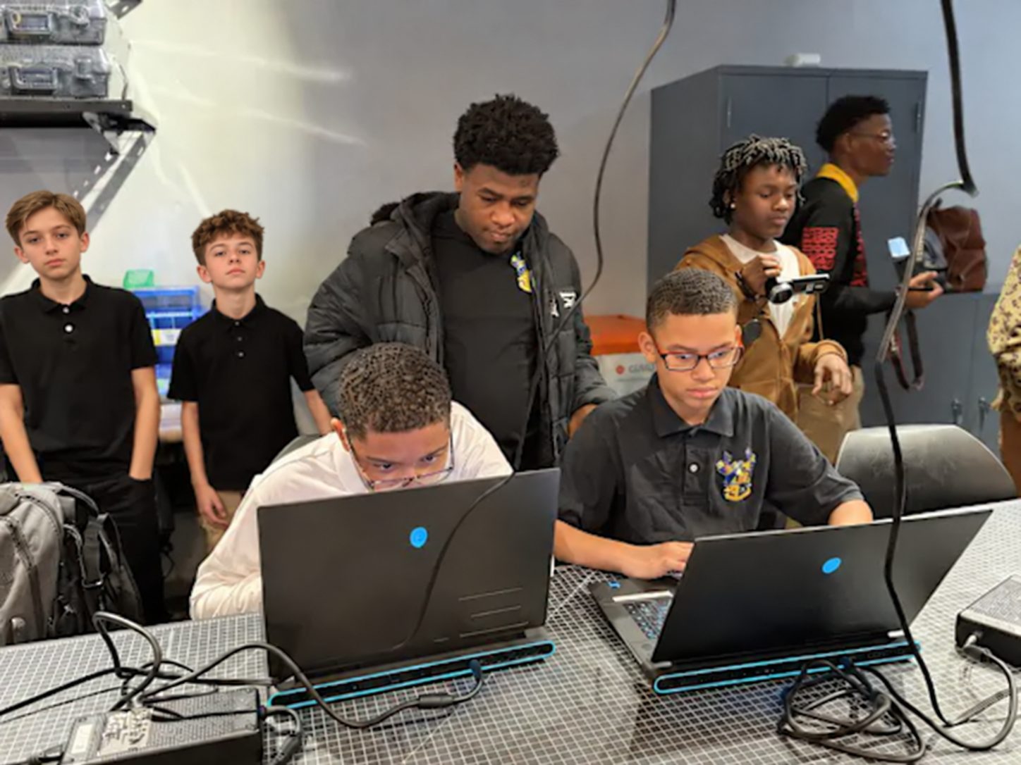Two middle school students working on laptops while a young adult mentor looks over their shoulders, with classmates standing behind them